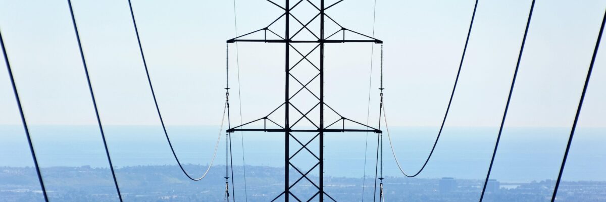 An image of transmission tower in the midground center, with draping transmission lines on either side extending from the foreground to the midground; in the background, a smoky valley in various shades of blue.