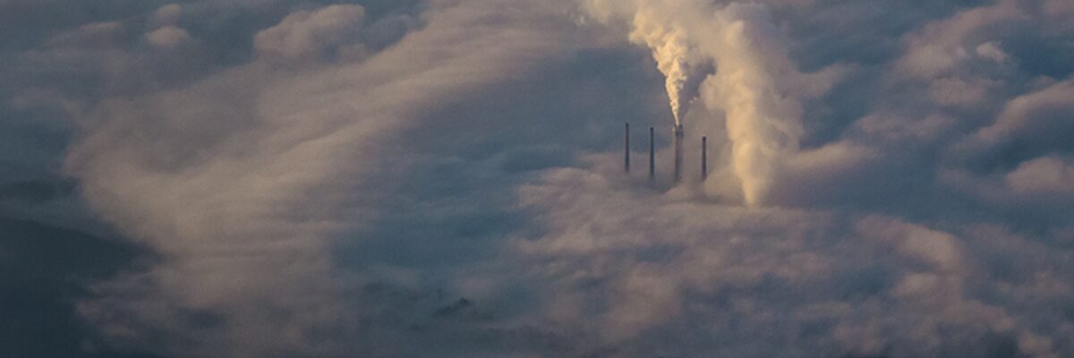 A birds-eye view of 4 smoke stacks, one releasing a large stream of pollution into the air around it.