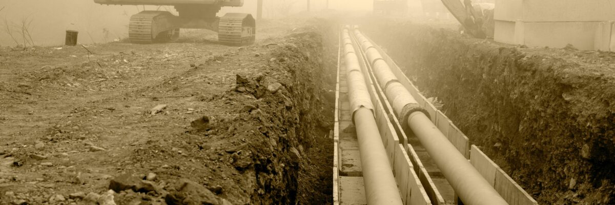 A construction vehicle reaching its digging arm over a pipeline, which is in the process of being buried in the ground.
