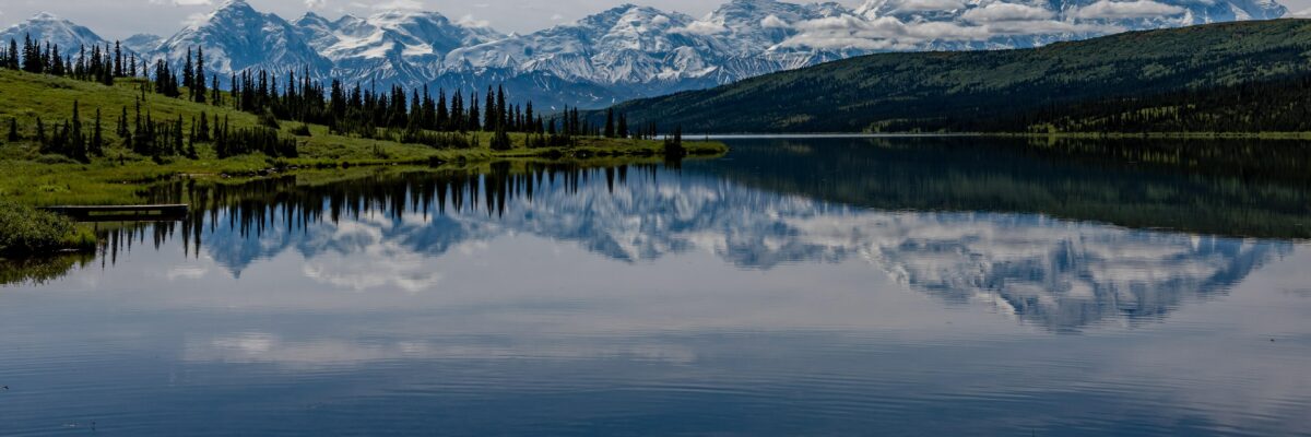 A clear blue lake reflects a scene of a mountain on a clear day; evergreen trees line the shore.
