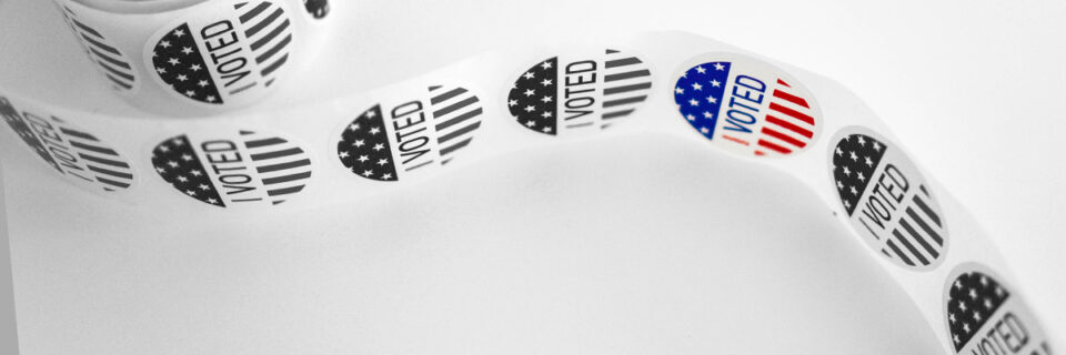 A roll of circular "I voted" stickers unfurled; the image is black and white except for one sticker, which is red and blue