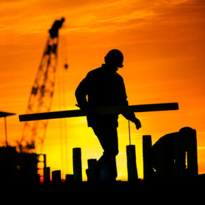 The black silhouette of a worker wearing a hard hat and carrying a beam contrasting against a burning orange sky; a crane looms in the distance.