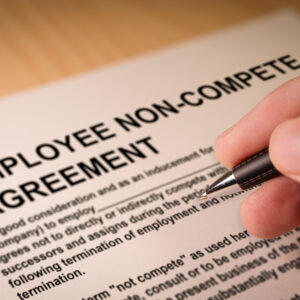 A person holding a pen over a contract titled "Employee Non-Compete Agreement"