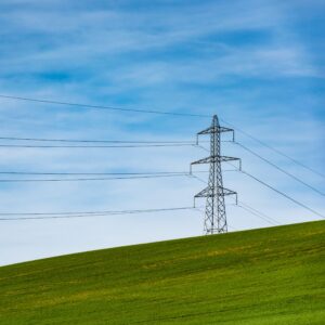 A transmission tower sits on a sloping green hill against a blue sky filled with wispy white clouds